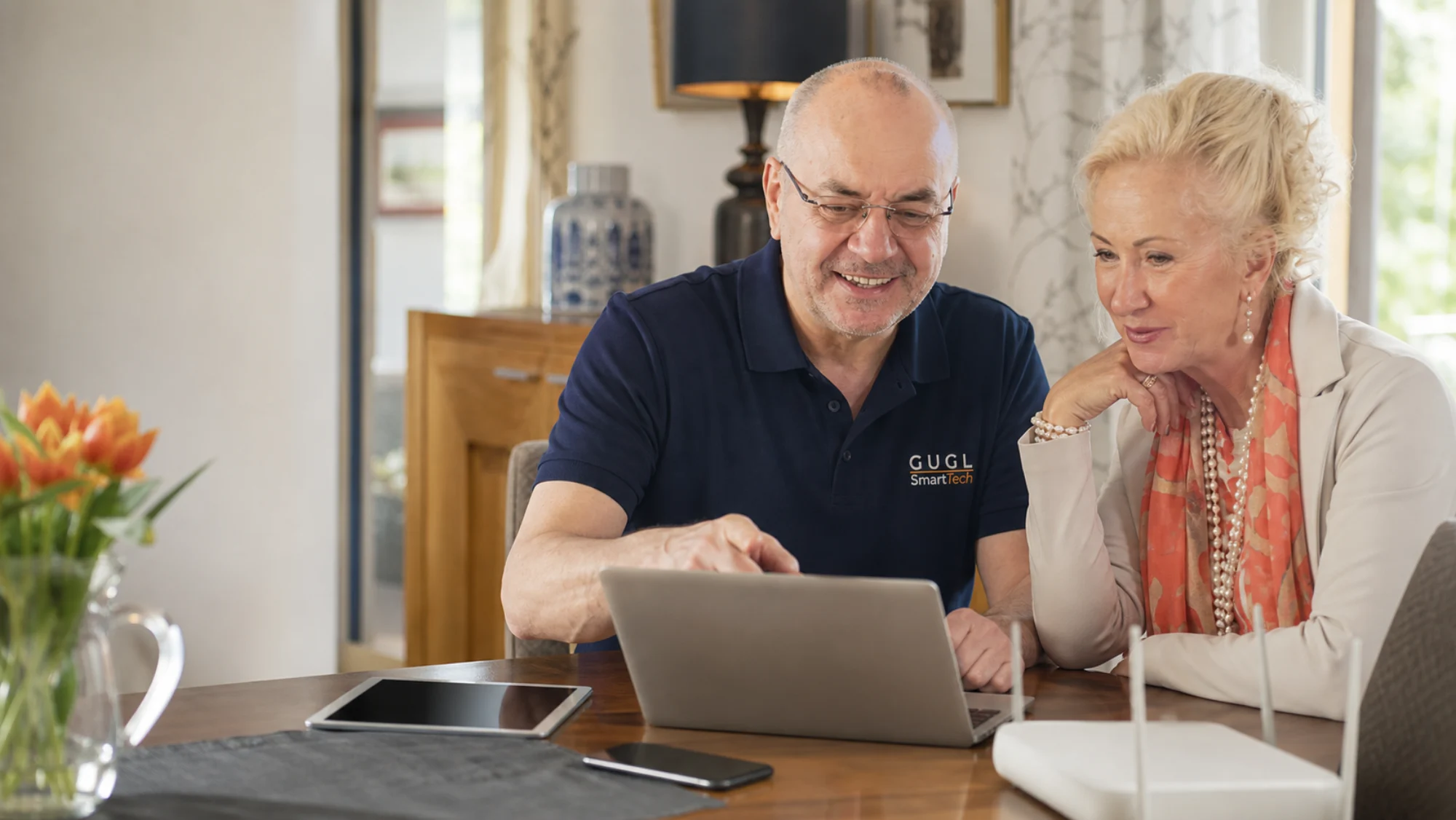 Günther Gugl erklärt einem älteren Ehepaar etwas am Laptop im Wohnzimmer